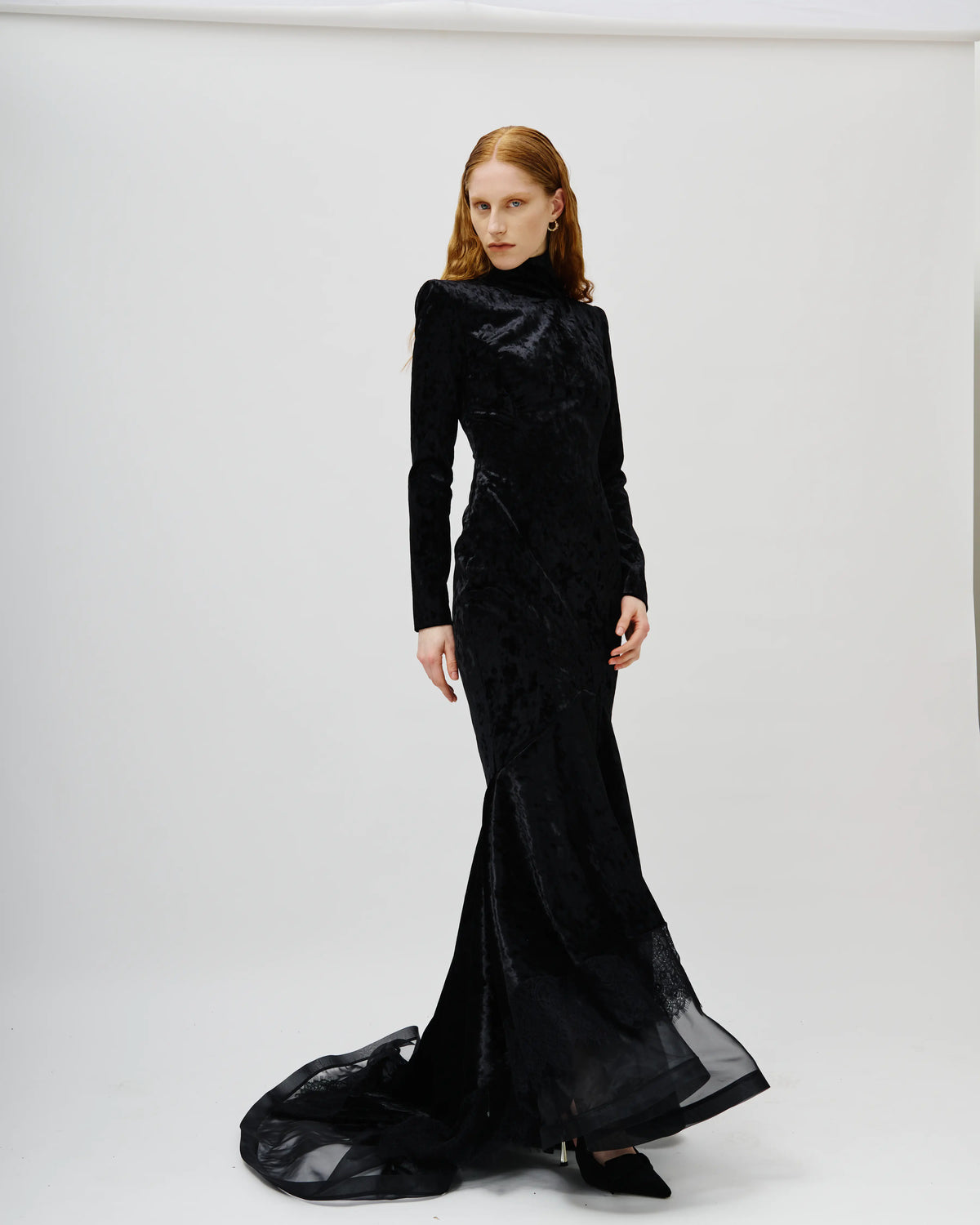 A model wearing a long-sleeved Avaro Figlio black velvet evening gown with a high mock neck and a dramatic train, photographed against a seamless white studio background.
