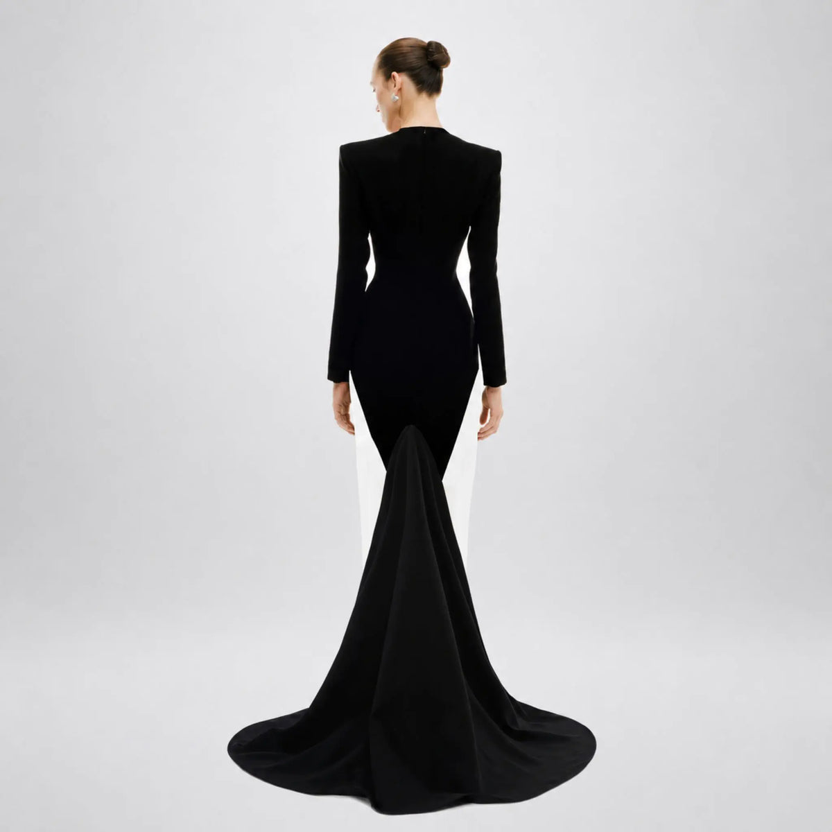 A woman viewed from the back wearing a fitted avaro figlio black long-sleeve evening gown with a dramatic train, photographed against a clean light gray studio background.