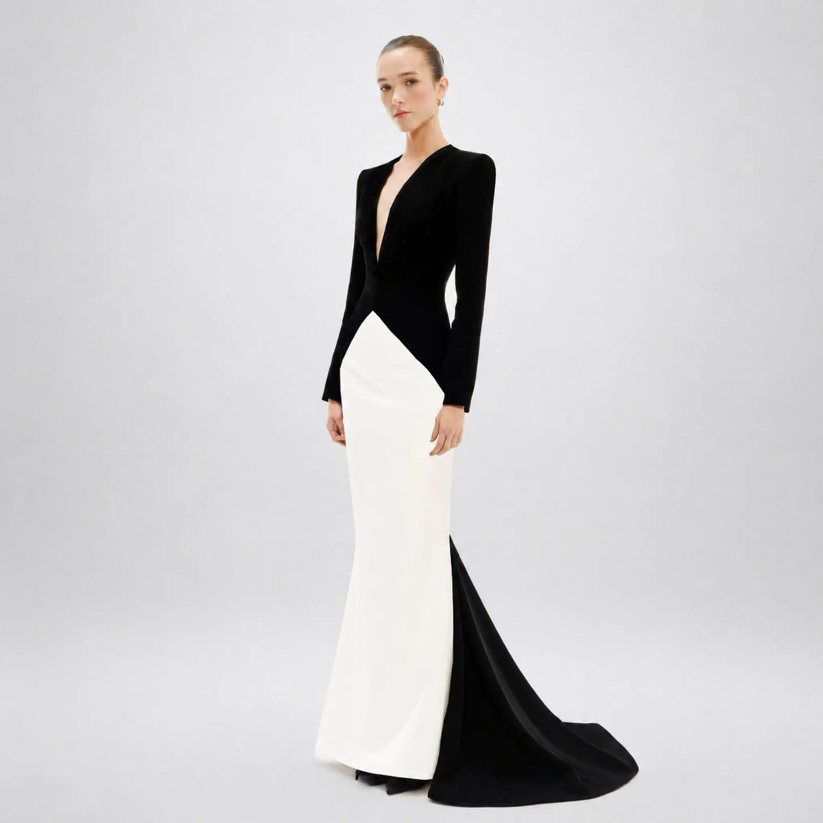 A model wears a long-sleeved floor-length avaro figlio gown with a color-blocked design, featuring a black velvet bodice and sleeves, a deep V-neckline, and a contrasting white skirt with a black train, set against a light gray studio background.