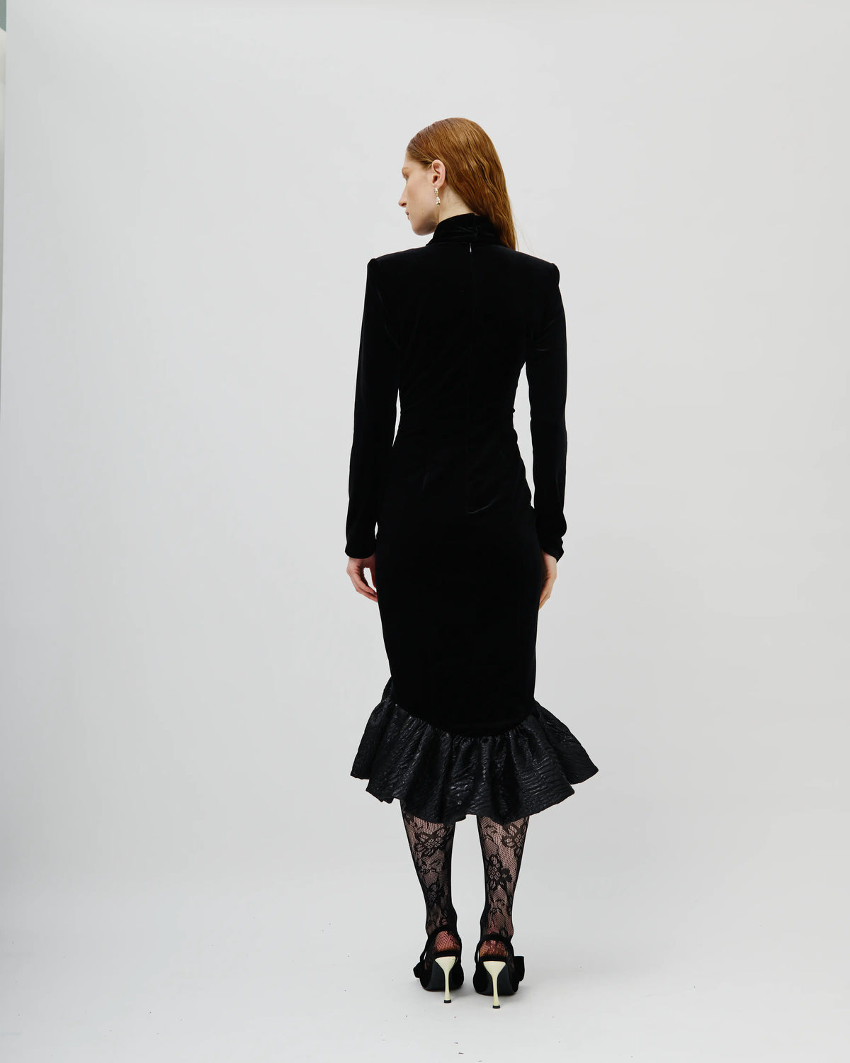 A model is shown from the back wearing a black Avaro Figlio velvet long-sleeve midi dress with structured shoulders and a ruffled hem, paired with black lace tights and black high heels, against a plain white studio background.