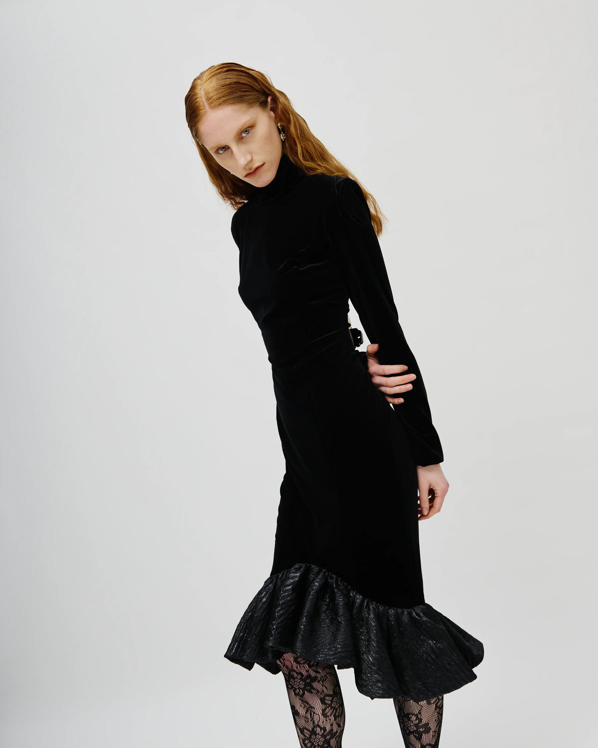 A model with red hair wearing a black Avaro Figlio velvet mock-neck long-sleeve midi dress with a ruffled lace hem, paired with black lace tights, against a light gray studio background.