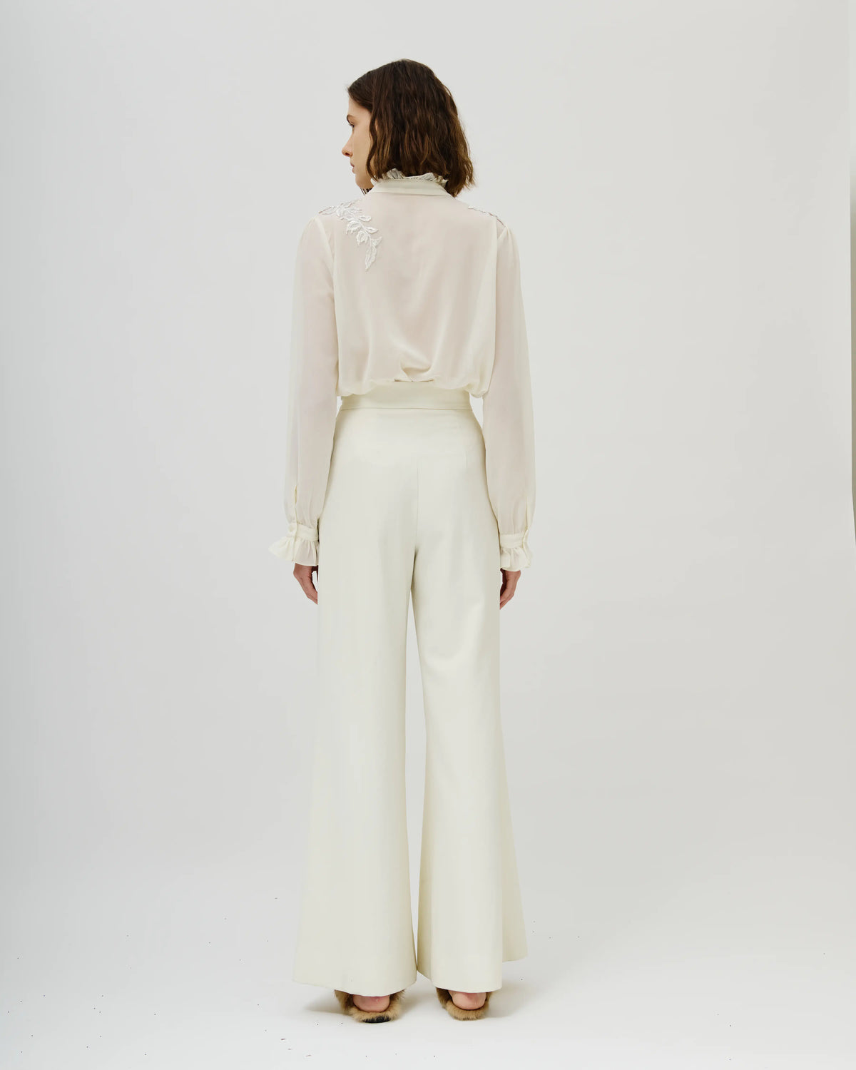 Model viewed from the back wearing a cream-colored Avaro Figlio long-sleeve blouse with delicate lace detailing on the shoulder and matching cream wide-leg trousers, set against a plain light gray studio background.
