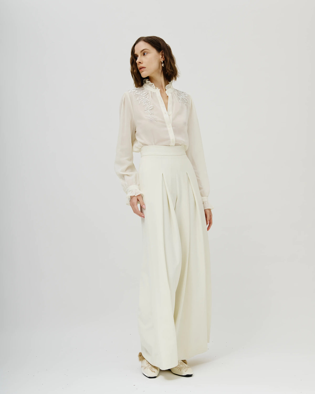 Model wearing a two-piece cream  Avaro Figlio consisting of a long-sleeve sheer blouse and matching wide-leg trousers, paired with flat tan sandals, against a plain light gray studio background.