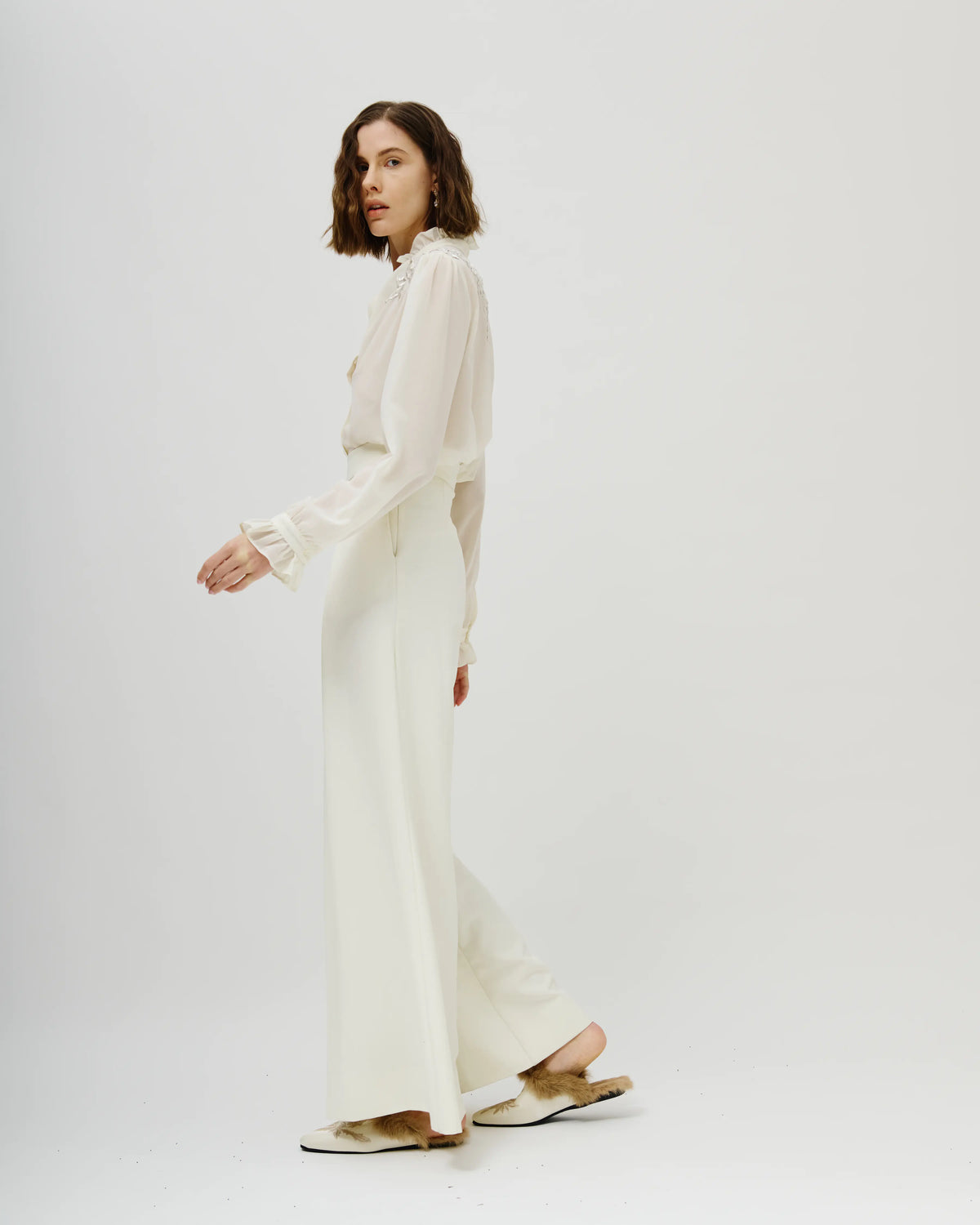 A woman modeling a two-piece cream  Avaro Figlio consisting of a long-sleeve sheer blouse and matching wide-leg trousers, paired with flat tan sandals, against a plain light gray studio background.