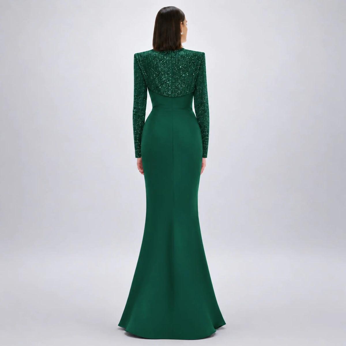 A woman viewed from the back wearing a floor-length avarofiglio forest green gown with a heavily embellished beaded back and long sleeves, photographed against a light gray studio background.