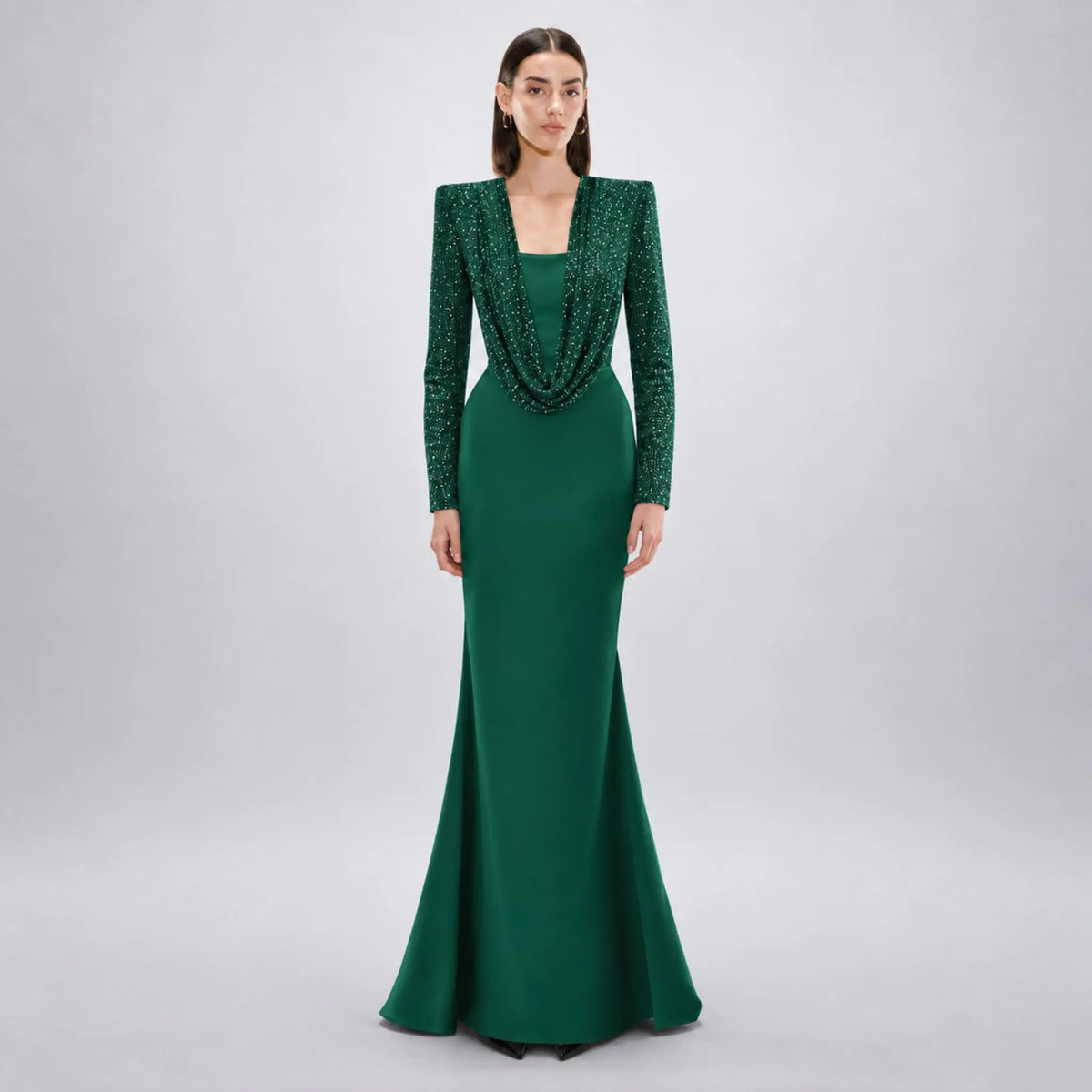 Model stands against a light gray studio background wearing a floor-length Avaro Figlio forest green gown with long sleeves, a draped cowl neckline, and a fitted mermaid silhouette.