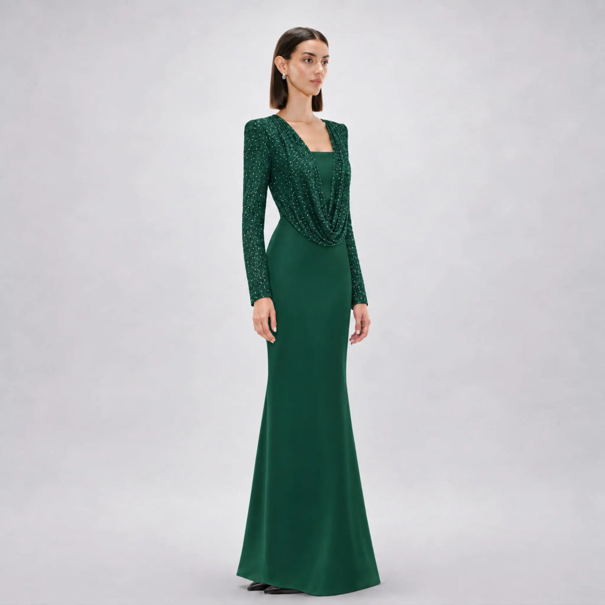 A woman modeling a floor-length avaro figlio forest green gown with long sleeves, a square neckline, and a draped cowl detail across the bodice, photographed against a light gray studio background.