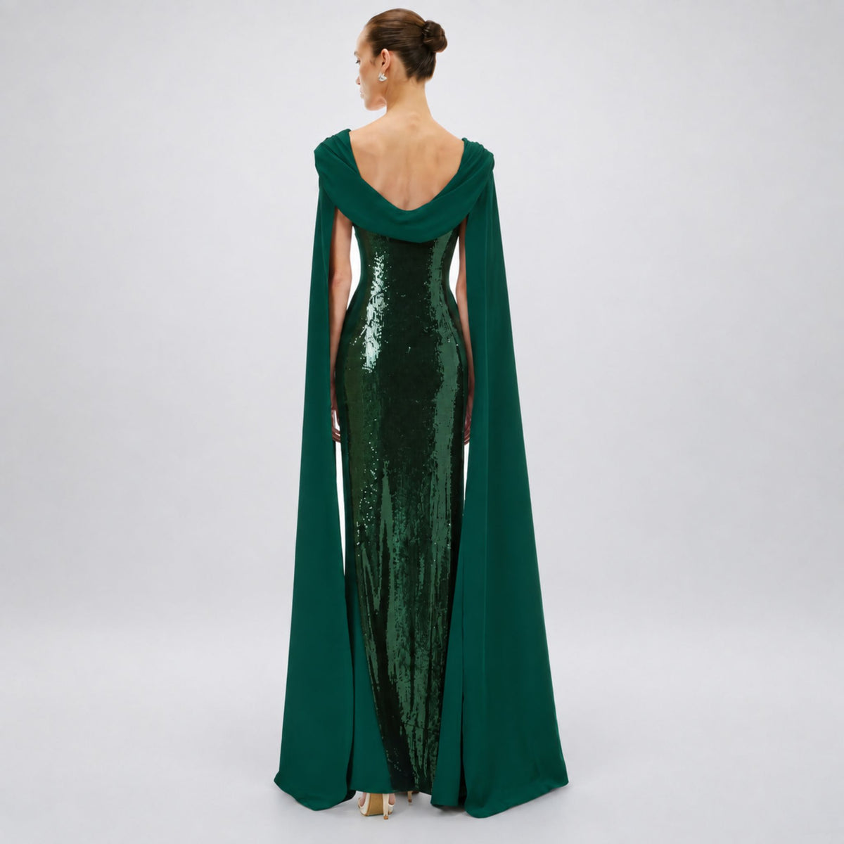 A woman viewed from the back wearing a floor-length Avaro Figlio forest green gown with a draped cape-like overlay and a fitted sequined bodice, photographed against a light gray studio background.