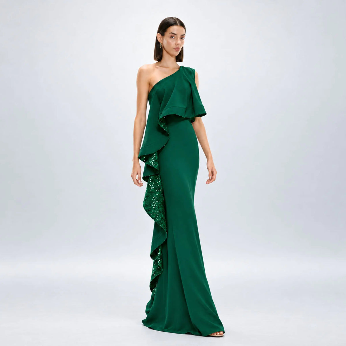 A woman modeling a floor-length one-shoulder avaro figlio forest green dress with cascading ruffles and sequined detailing on a light gray studio background.