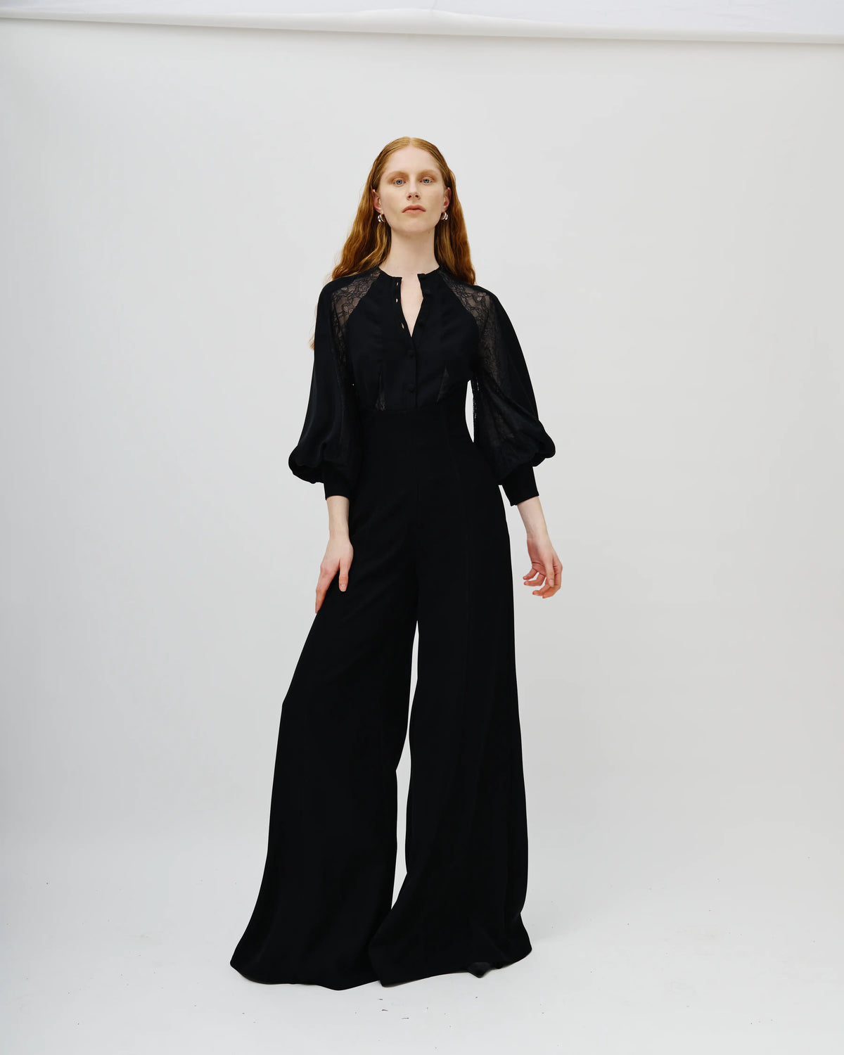A woman modeling a black Avaro Figlio wide-leg jumpsuit with sheer lace shoulder panels and balloon sleeves, standing against a plain light gray studio background.