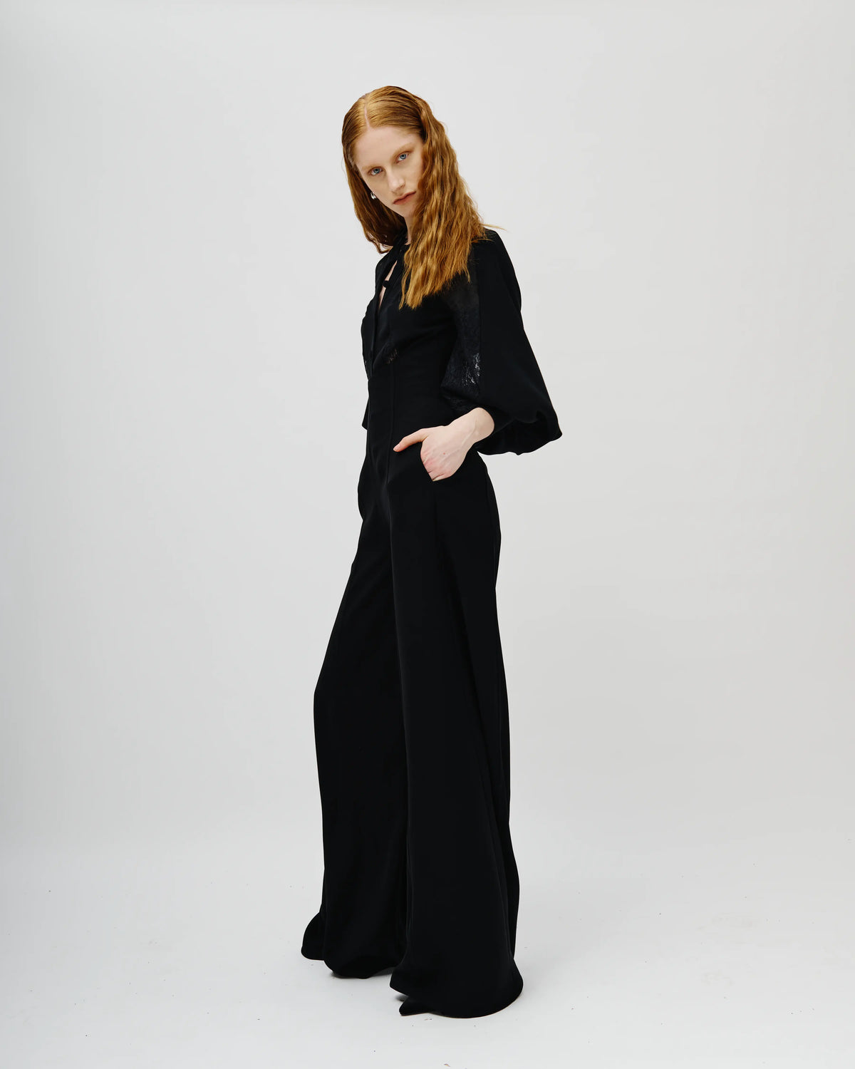 A model with long red hair wearing a black Avaro Figlio wide-leg jumpsuit with three-quarter sleeves and a high collar, standing against a plain light gray studio background.
