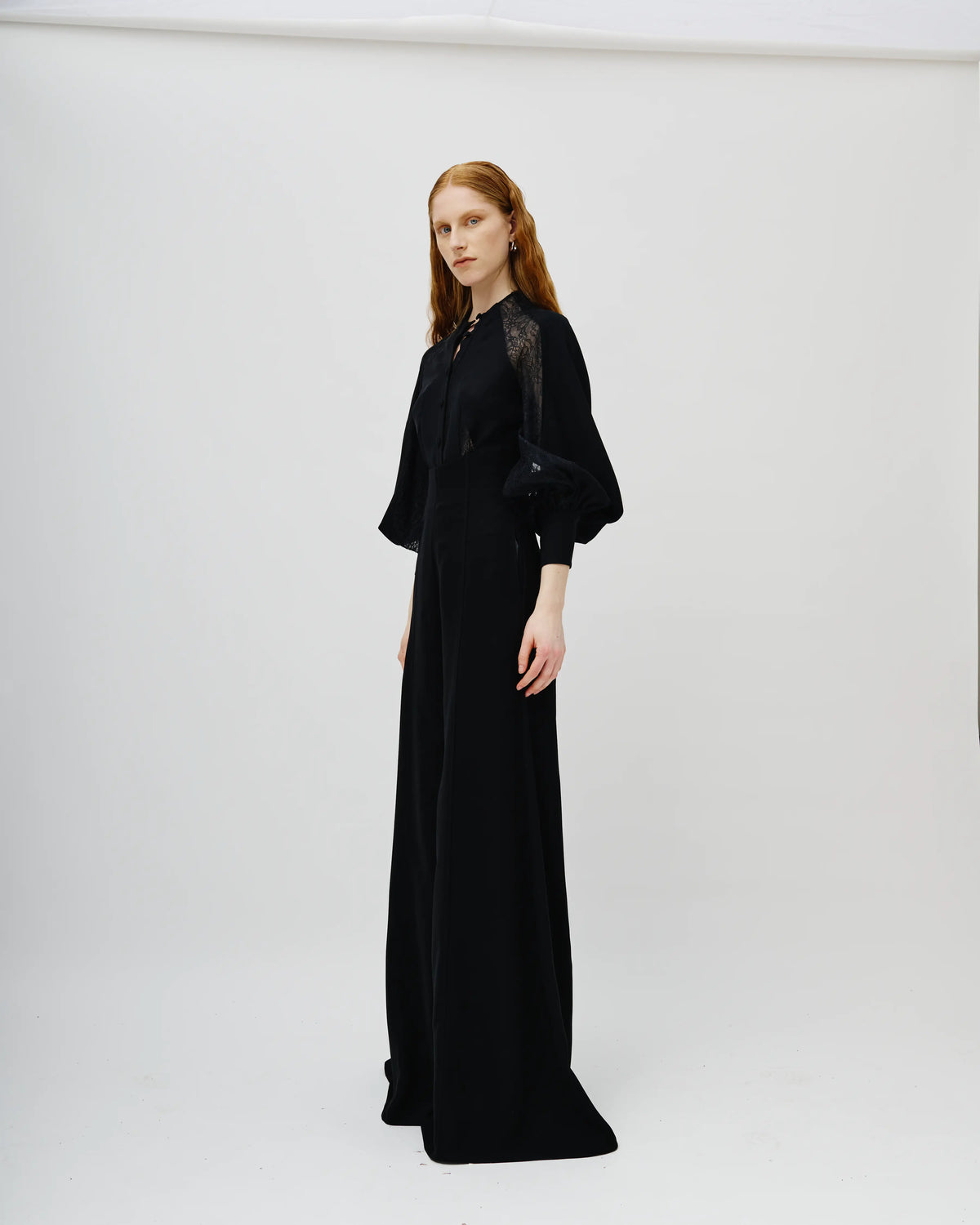 A model with red hair wearing a long black Avaro Figlio dress with sheer black lace sleeves, standing against a plain white studio background.