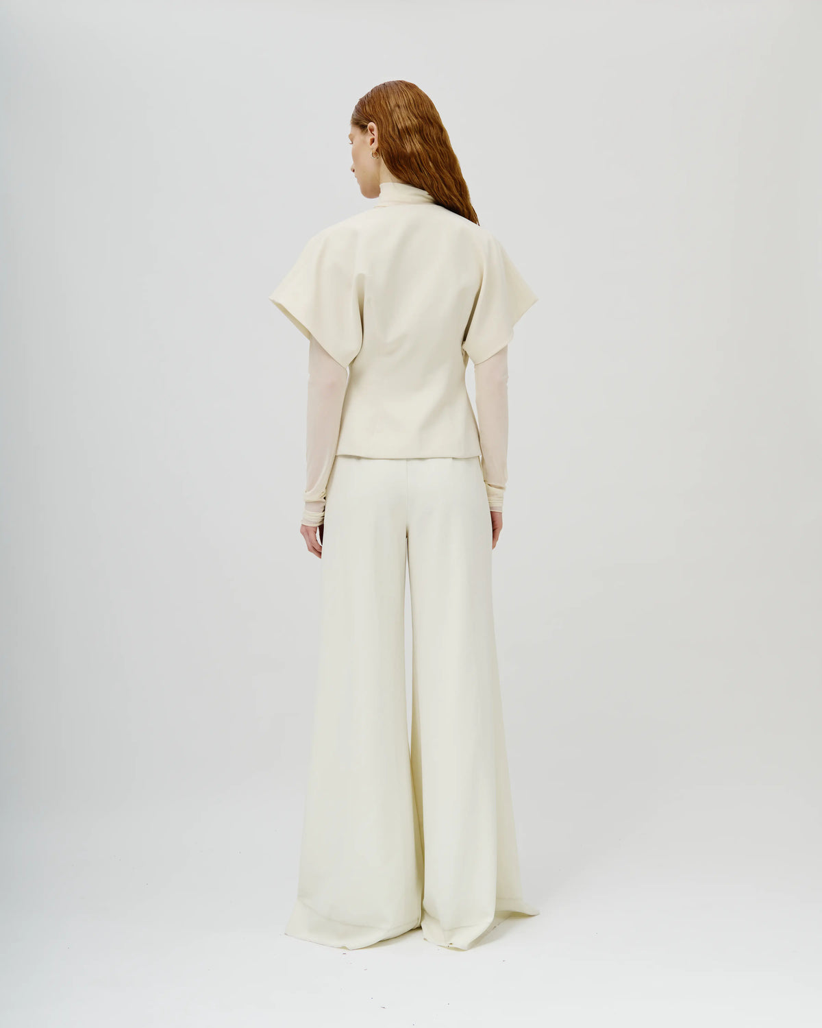 Model viewed from the back wearing a cream-colored Avaro Figlio two-piece outfit consisting of a short-sleeved cropped top with sheer long sleeves and matching wide-leg trousers, set against a plain light gray studio background.