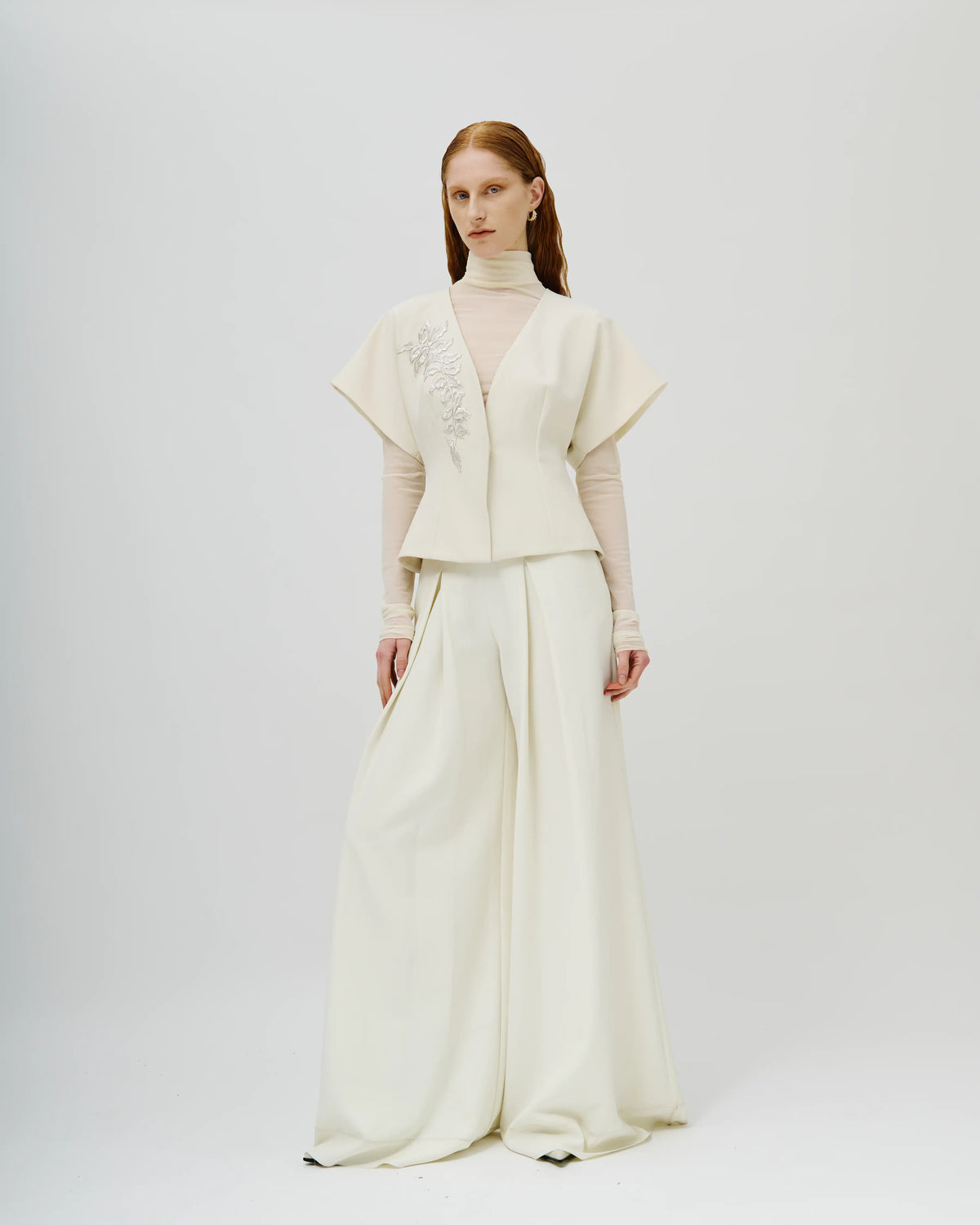 Model stands against a light gray studio background wearing a cream-colored Avaro Figlio two-piece outfit consisting of a short-sleeved cropped jacket with floral embroidery on the chest and matching wide-leg trousers.