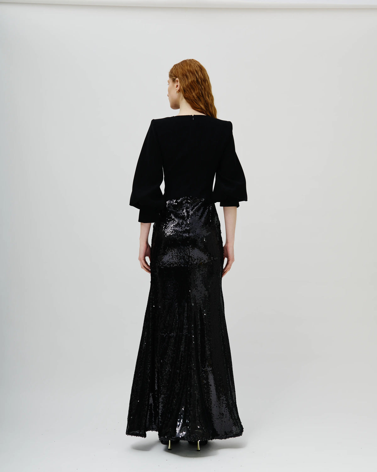 A model viewed from the back wearing an Avaro Figlio black velvet top with three-quarter puff sleeves and a high-waisted, floor-length black sequined skirt, set against a plain light gray studio background.
