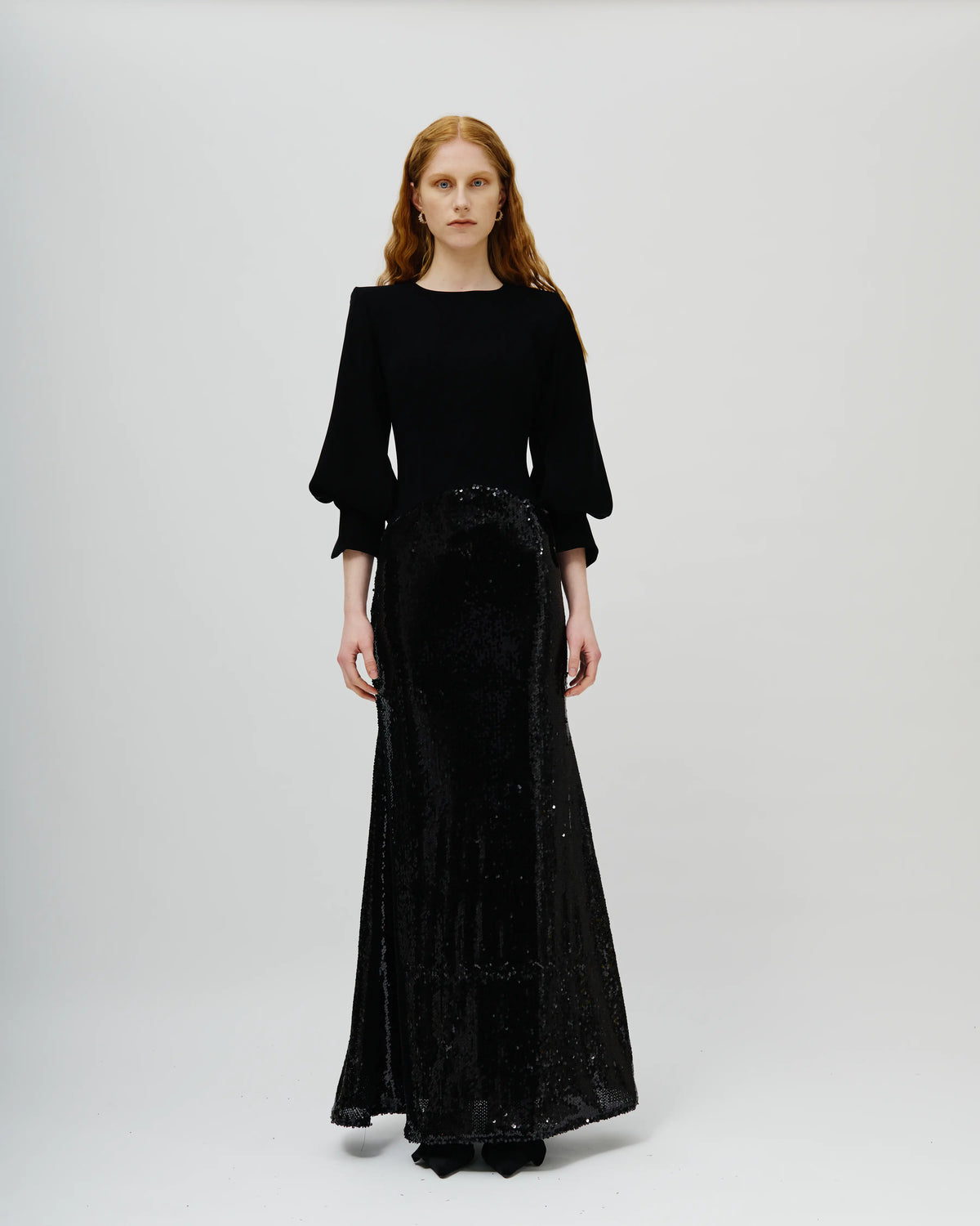 A model wearing a long black dress with a solid black long-sleeve top and a heavily embellished, sequined black skirt, standing against a light gray studio background.