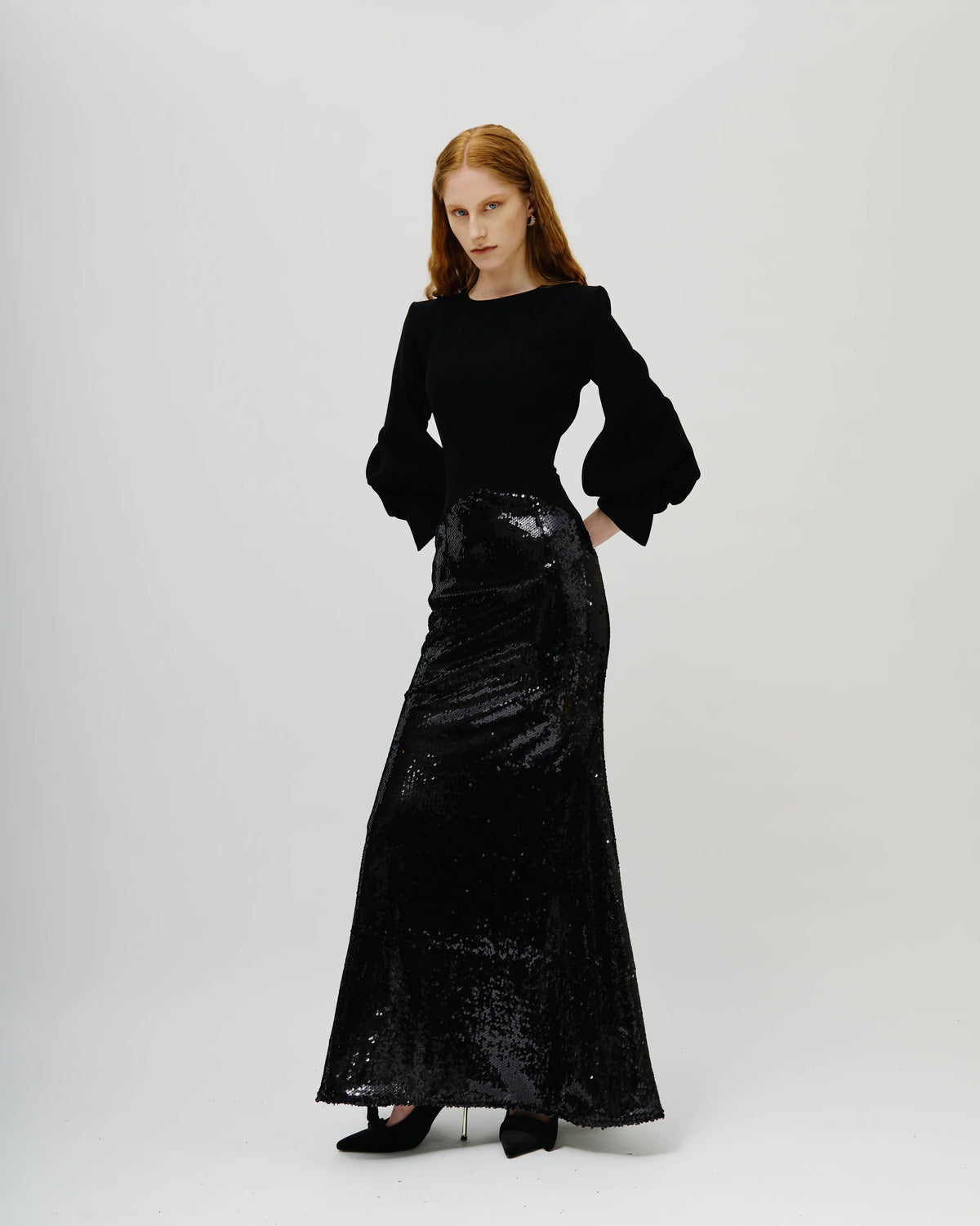 A model wearing an Avaro Figlio long black dress with a solid black long-sleeve top and a heavily embellished, sequined black skirt, standing against a light gray studio background.