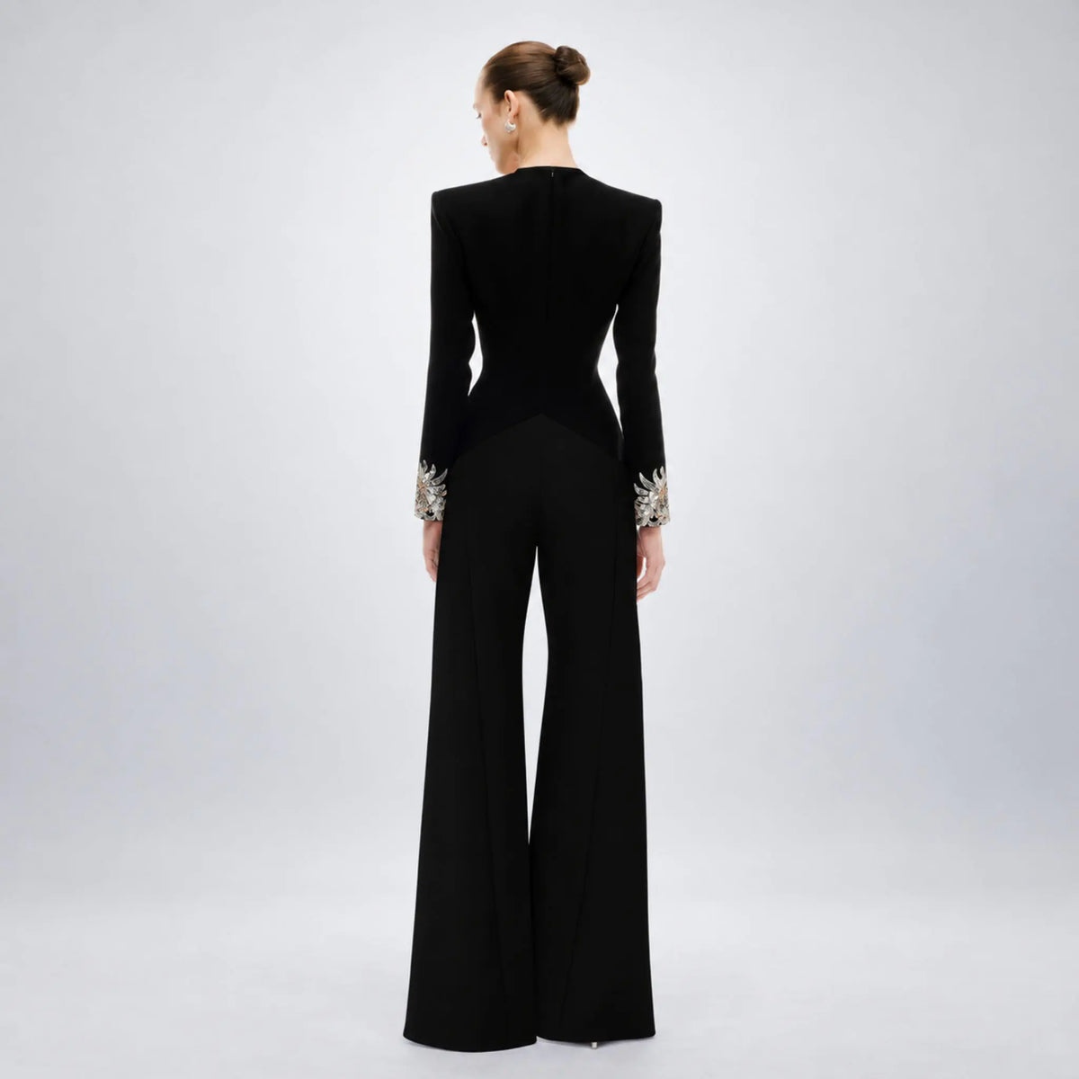 A woman stands with her back to the camera wearing a black avarofiglio long-sleeve jumpsuit with structured shoulders and ornate silver embellishments on the cuffs, photographed against a light gray studio background.