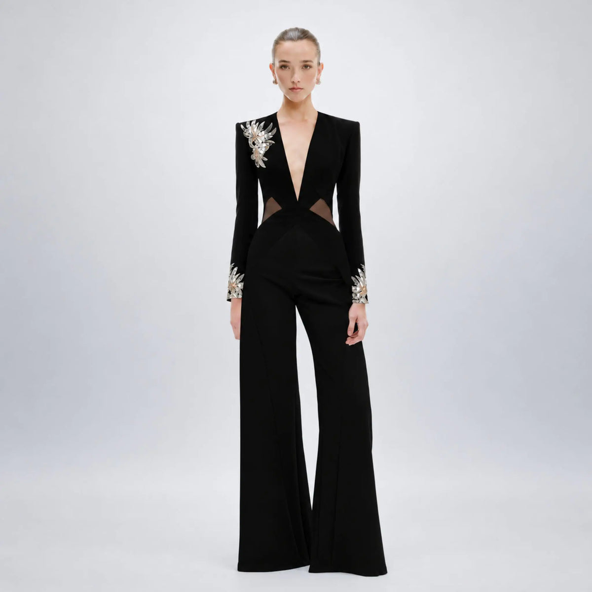 A model wears a black Avaro Figlio long-sleeve wide-leg jumpsuit with dramatic silver and white floral embellishments on the shoulders and cuffs, set against a light gray studio background.