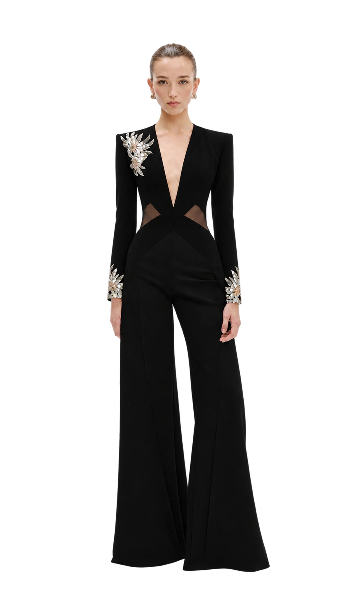 A model wears a black Avaro Figlio long-sleeve wide-leg jumpsuit with dramatic silver and white floral embellishments on the shoulders and cuffs, set against a light gray studio background.