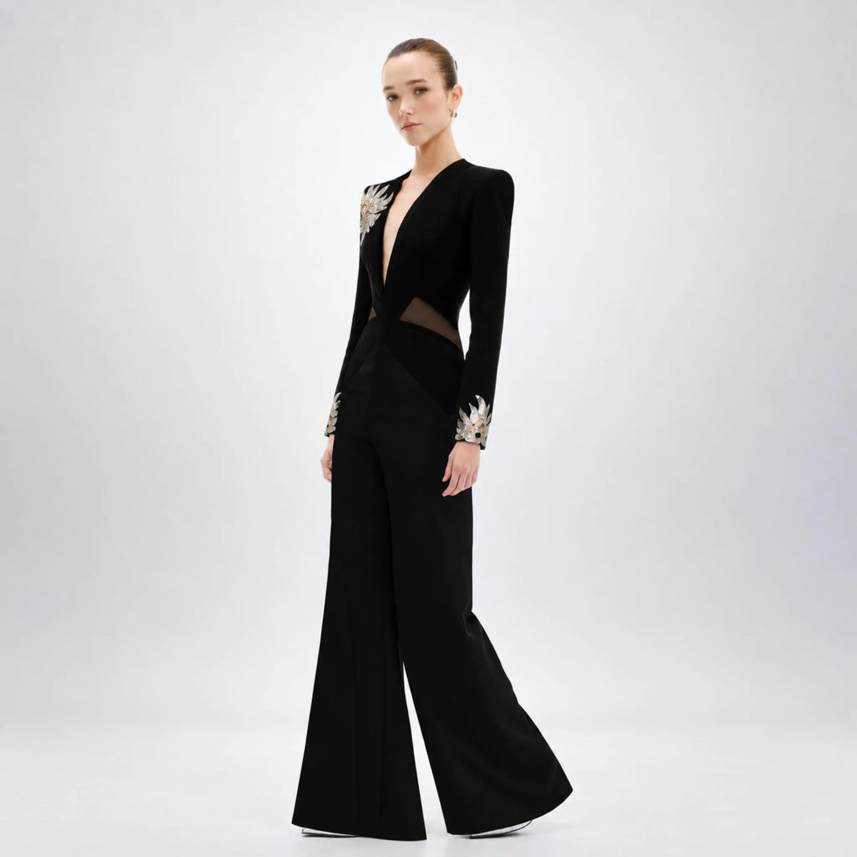 A model wearing a black long-sleeve AvaroFiglio jumpsuit with sheer mesh panels and ornate silver embellishments on the shoulders and cuffs, set against a plain light gray studio background.