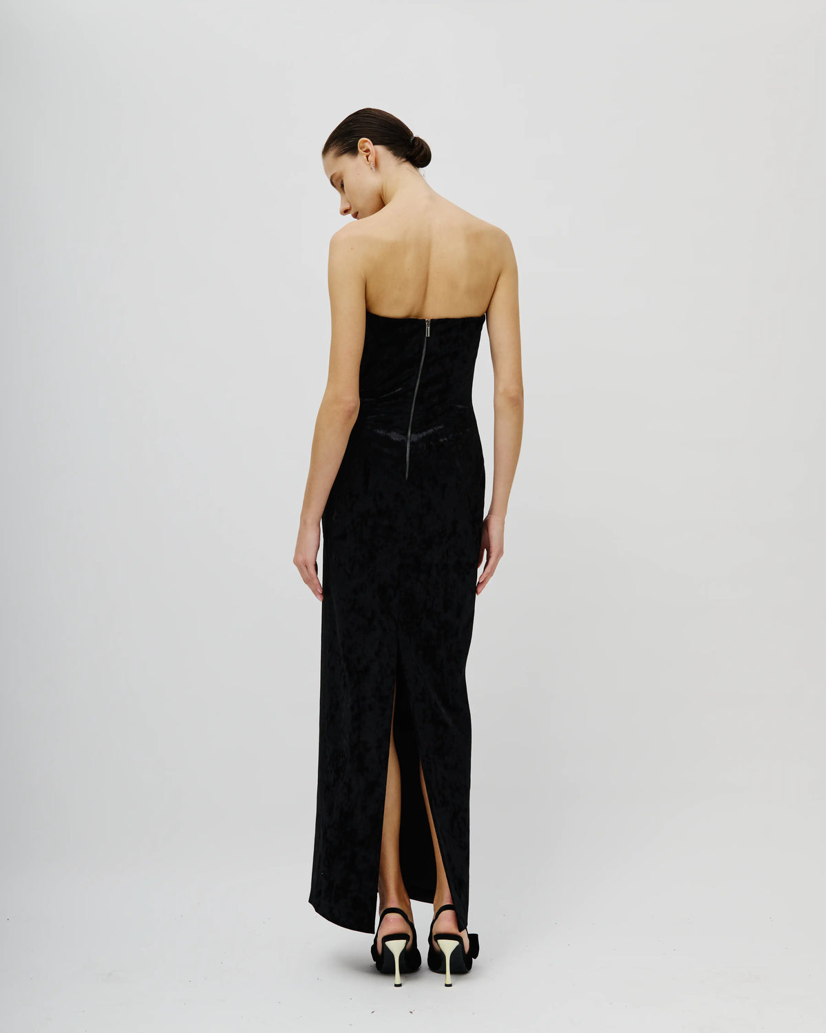 A woman modeling an Avaro Figlio strapless black velvet maxi dress from the back, featuring a structured corset-style waist, a high front slit, and black high-heeled sandals, set against a plain light gray studio background.