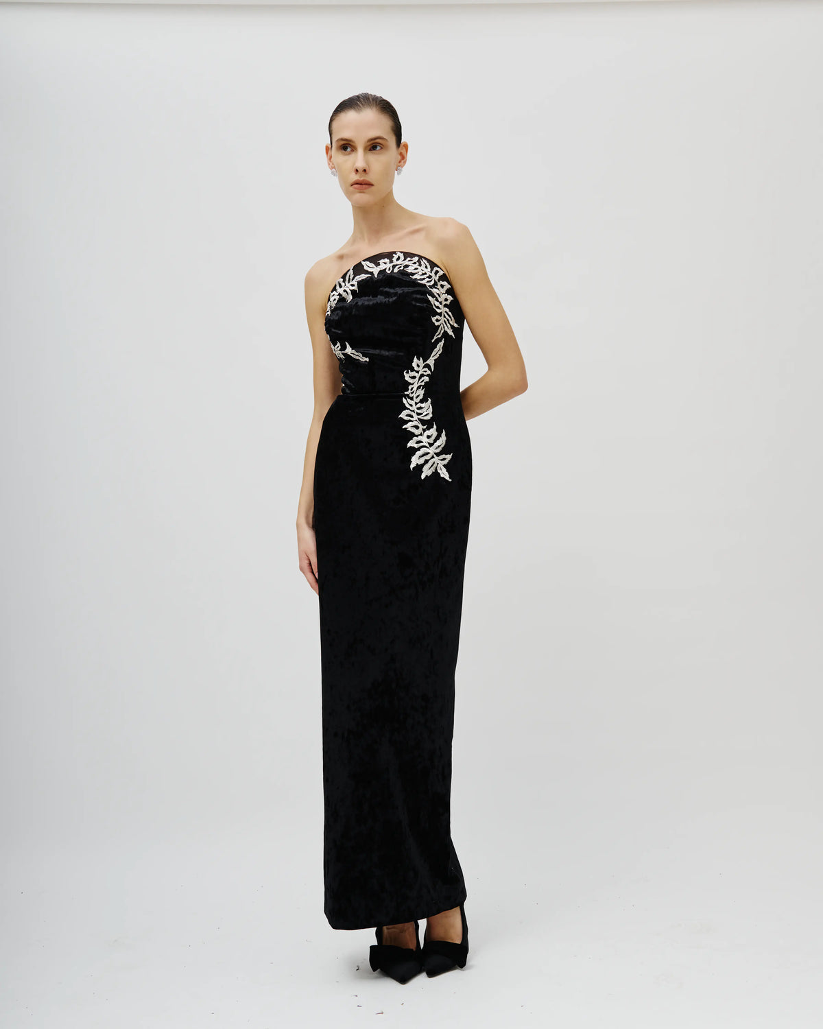 A model stands against a light gray studio background wearing an Avaro Figlio strapless black velvet gown with silver floral embroidery on the bodice and cascading down the left side of the skirt.
