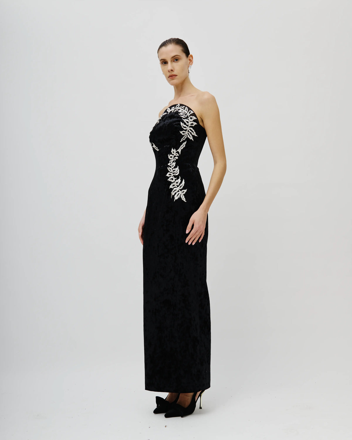 A woman modeling a strapless black Avaro Figlio velvet maxi gown with white floral embroidery on the bodice and side, paired with black pointed-toe heels, against a light gray studio background.