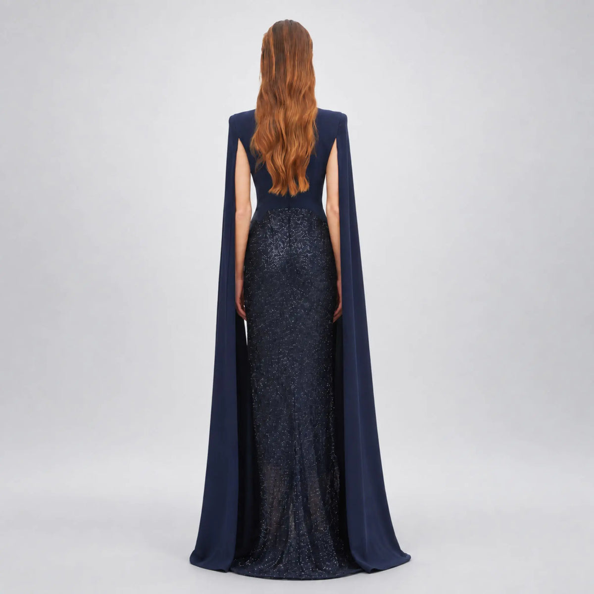 A woman viewed from the back wearing a floor-length Avaro Figlio navy gown with a fitted sequined skirt, a draped cape-like back overlay, and a long wavy auburn hair, photographed against a light gray studio background.