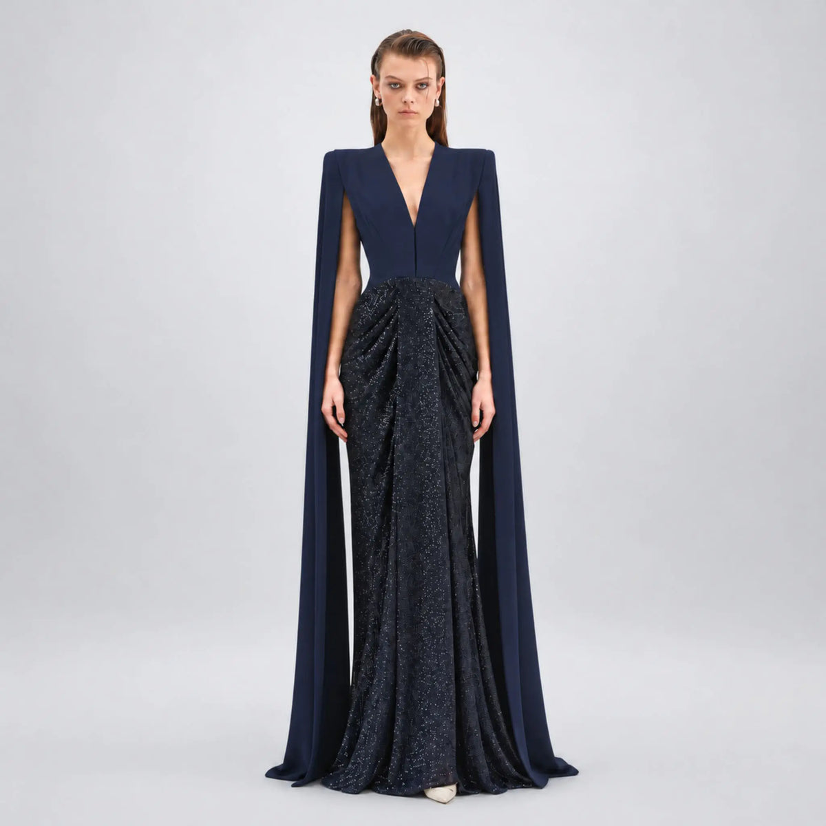 A model stands against a light gray studio background wearing a floor-length Avaro Figlio gown with a deep V-neckline, structured cap sleeves, and a long flowing cape. The bodice is a solid dark navy, while the skirt is a shimmering dark blue-gray textured fabric with a draped, gathered silhouette.