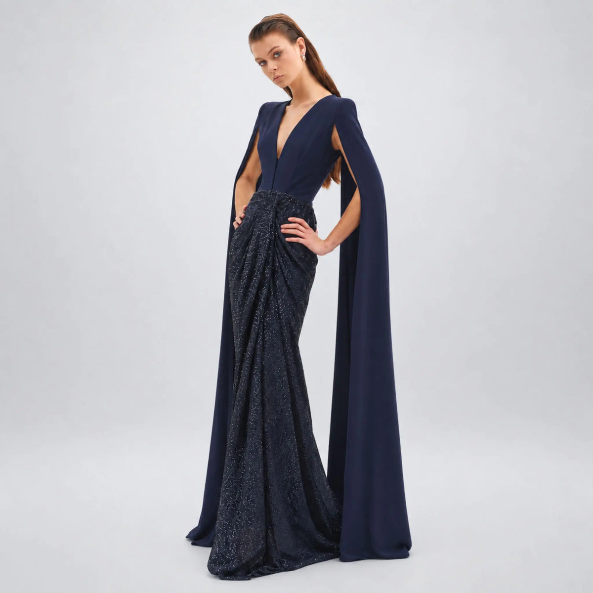 A model wearing a floor-length Avaro Figlio navy blue evening gown with a draped cape-like sleeve, deep V-neckline, and textured beaded skirt, photographed against a light gray studio background.