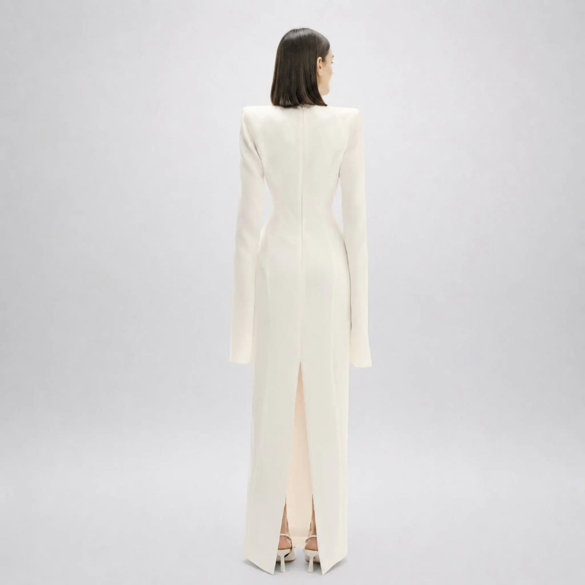 A model viewed from the back wearing a floor-length, structured off-white Avaro Figlio gown with long sleeves and a high front slit, set against a light gray studio background.