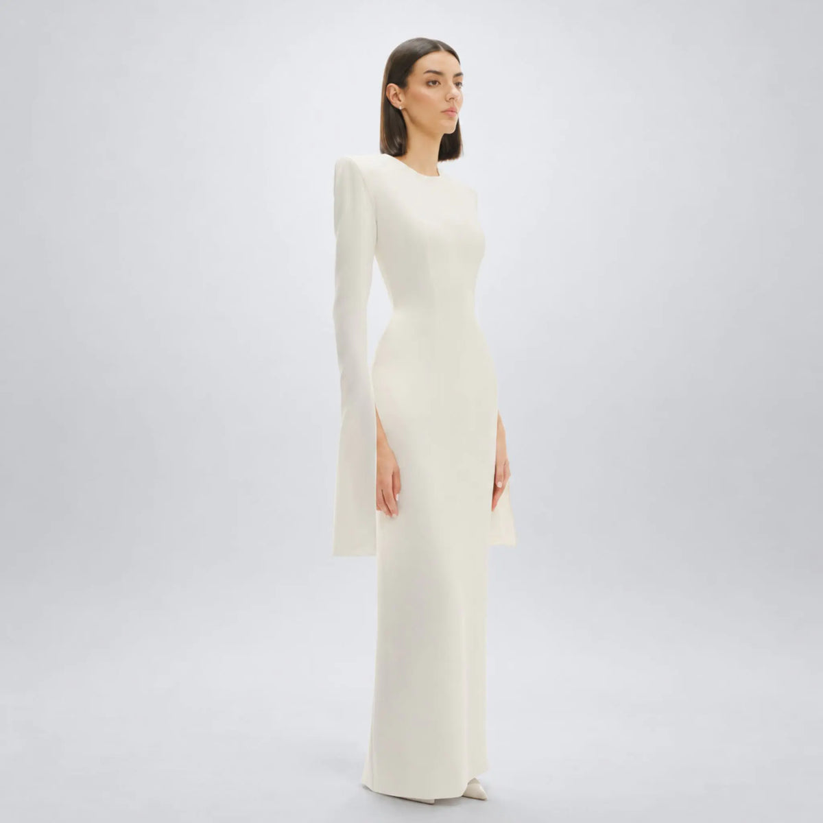 A woman modeling a floor-length white avaro-figlio gown with a long flared sleeves, and a fitted silhouette, photographed against a clean light gray studio background.