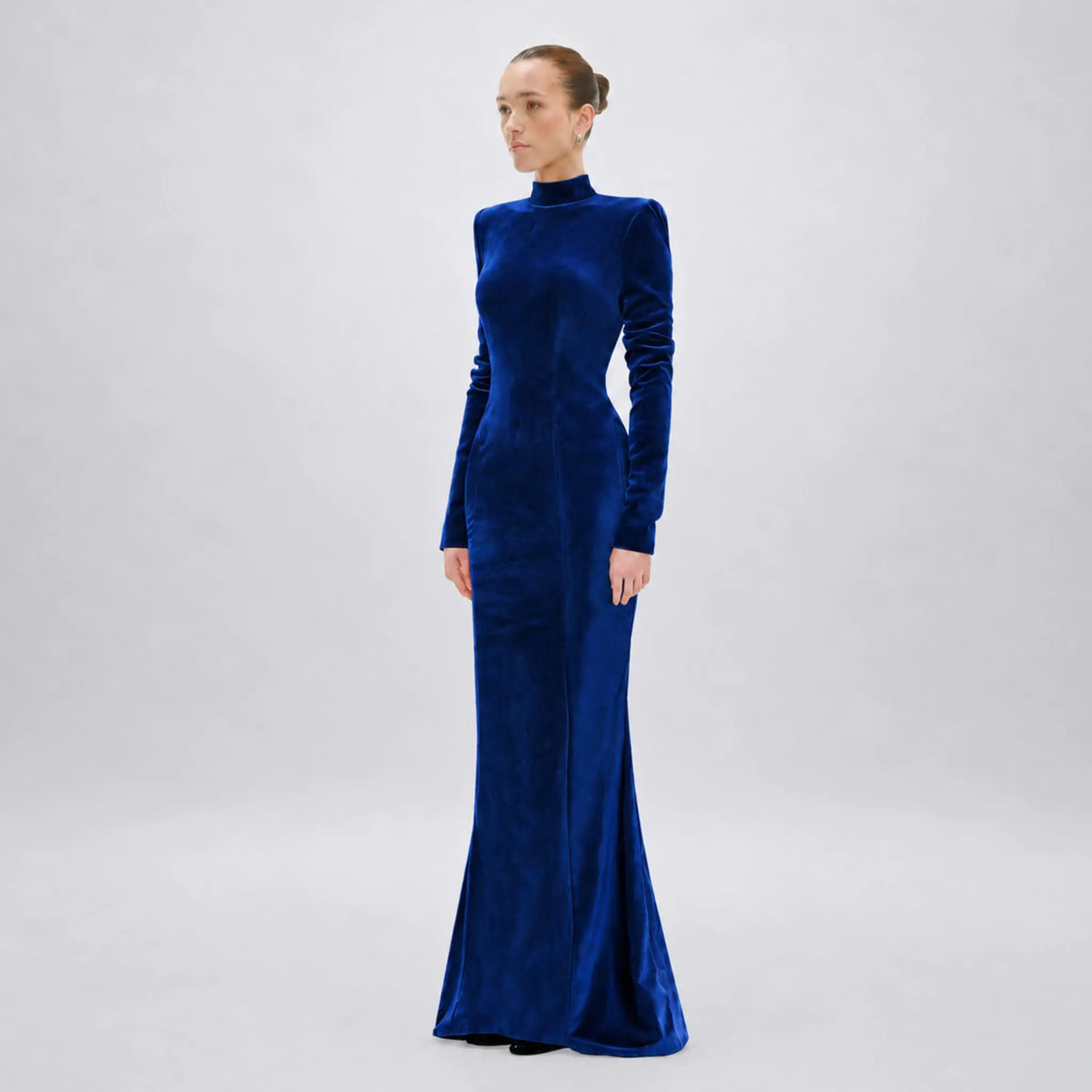 A woman modeling a long-sleeved, high-neck floor-length avaro figlio gown in rich royal blue velvet against a light gray studio background.