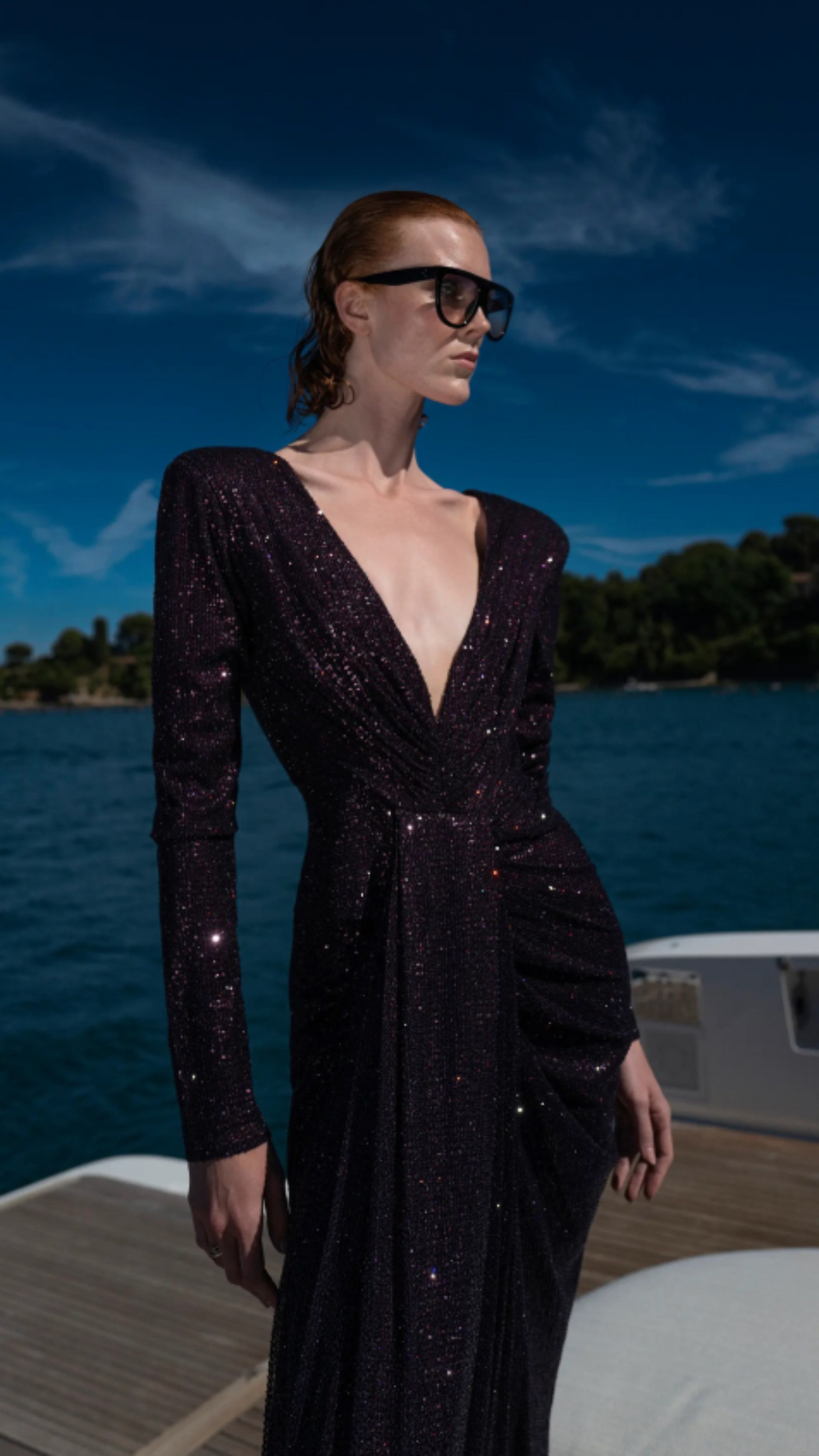 A woman wearing a sparkling long-sleeve wrap-style maxi dress in deep black or very dark purple, standing on a yacht deck with blue water and sky in the background.