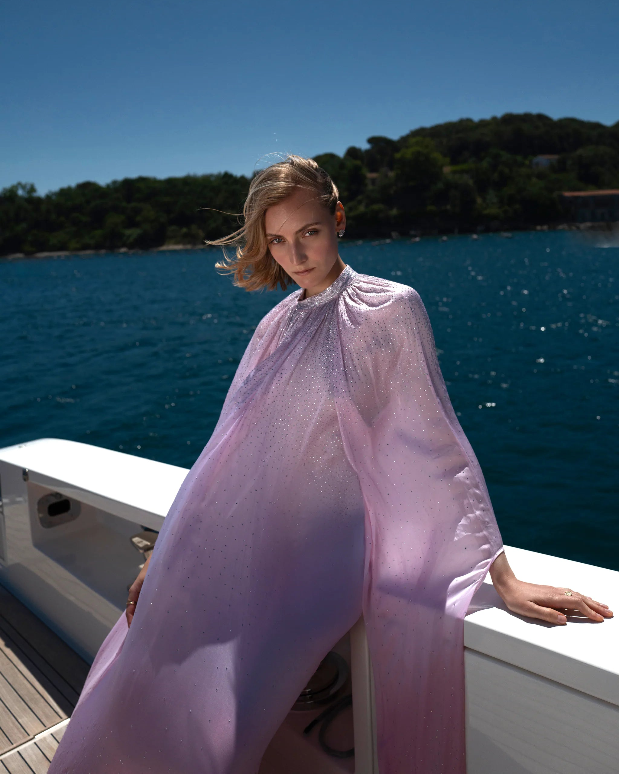 A woman wearing a flowing sheer lavender cape stands on a boat deck with blue water and a tree-lined shore in the background.