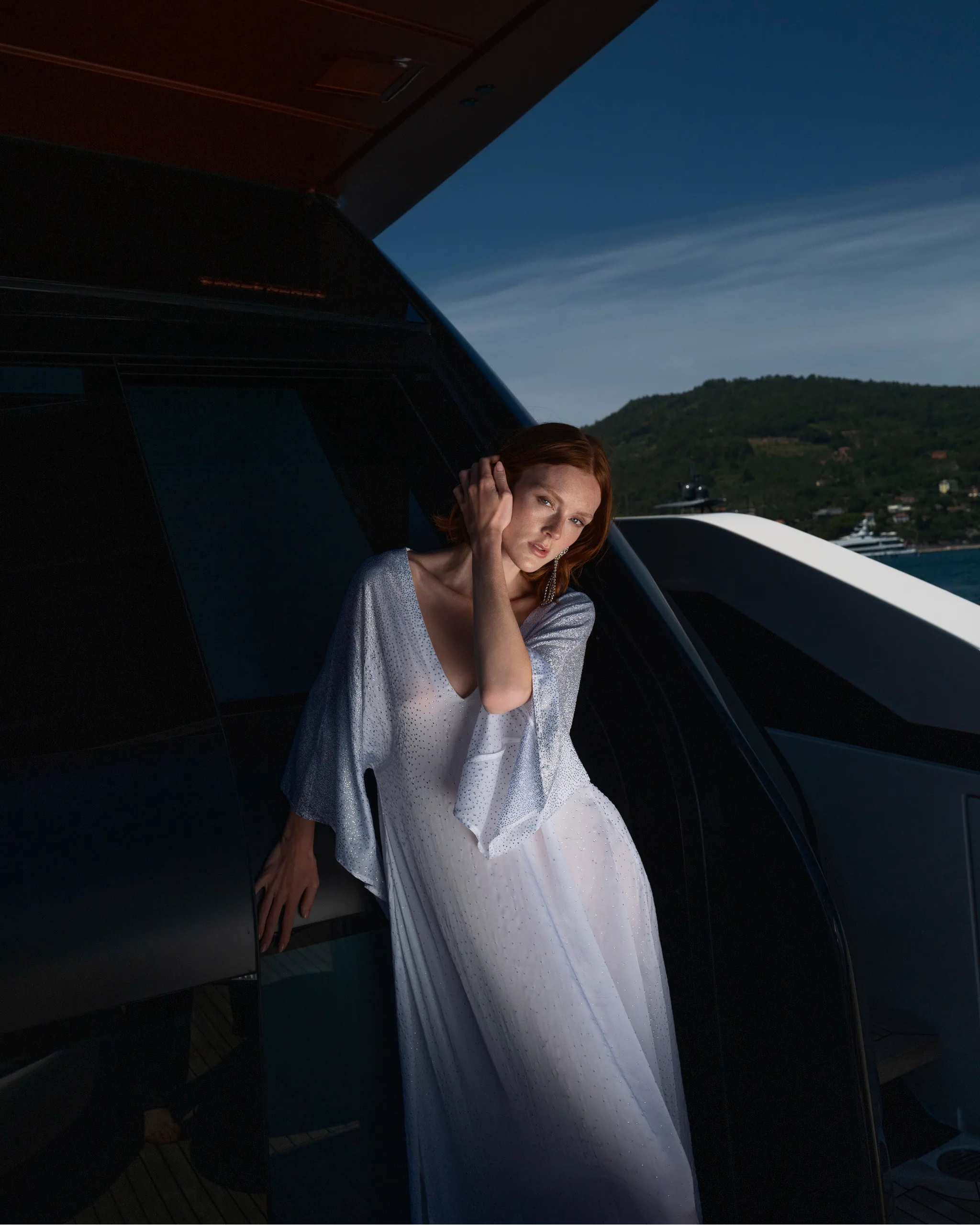 A woman in a flowing white dress leans against the open doorway of a yacht on a blue waterway with hills in the background.
