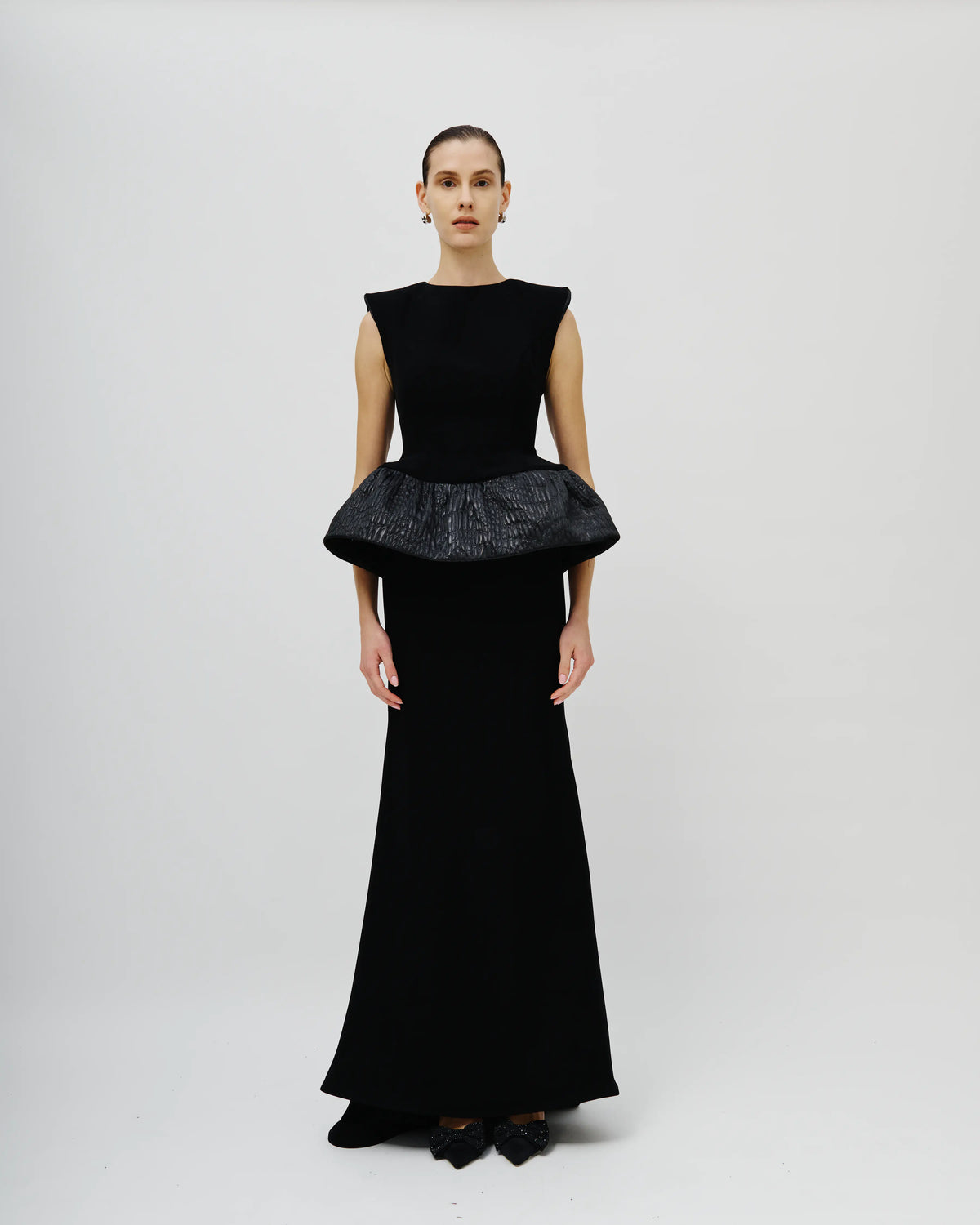 A model stands against a light gray studio background wearing a floor-length Avaro Figlio black gown with a structured peplum waist and a textured, patterned fabric on the waistband.