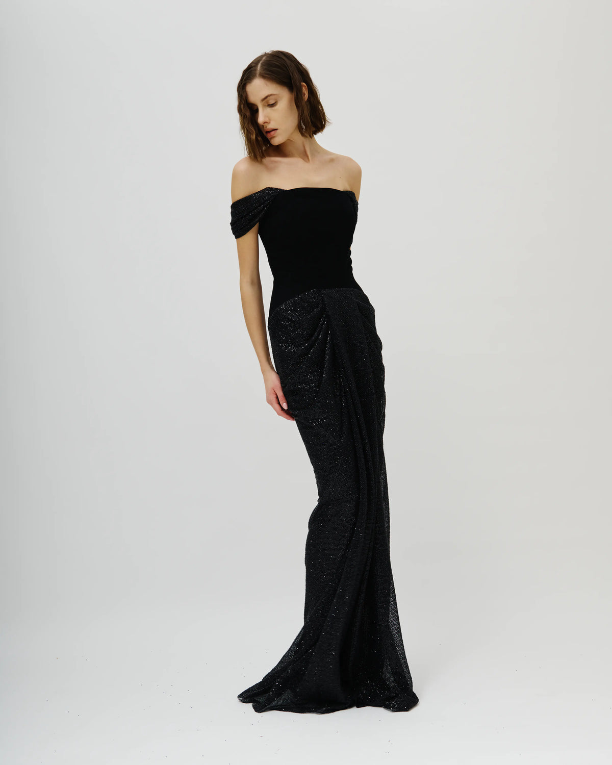 A woman modeling a floor-length Avaro Figlio black off-the-shoulder gown with a structured bodice and a draped, textured skirt, photographed against a plain light gray studio background.