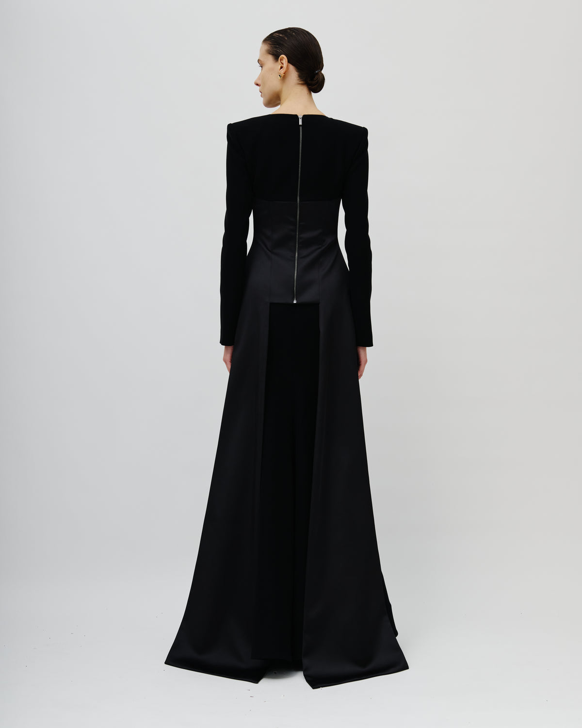 A model shown from the back wearing a long black Avaro Figlio evening gown with structured shoulders, long sleeves, and a front zipper detail, photographed against a light gray studio background.