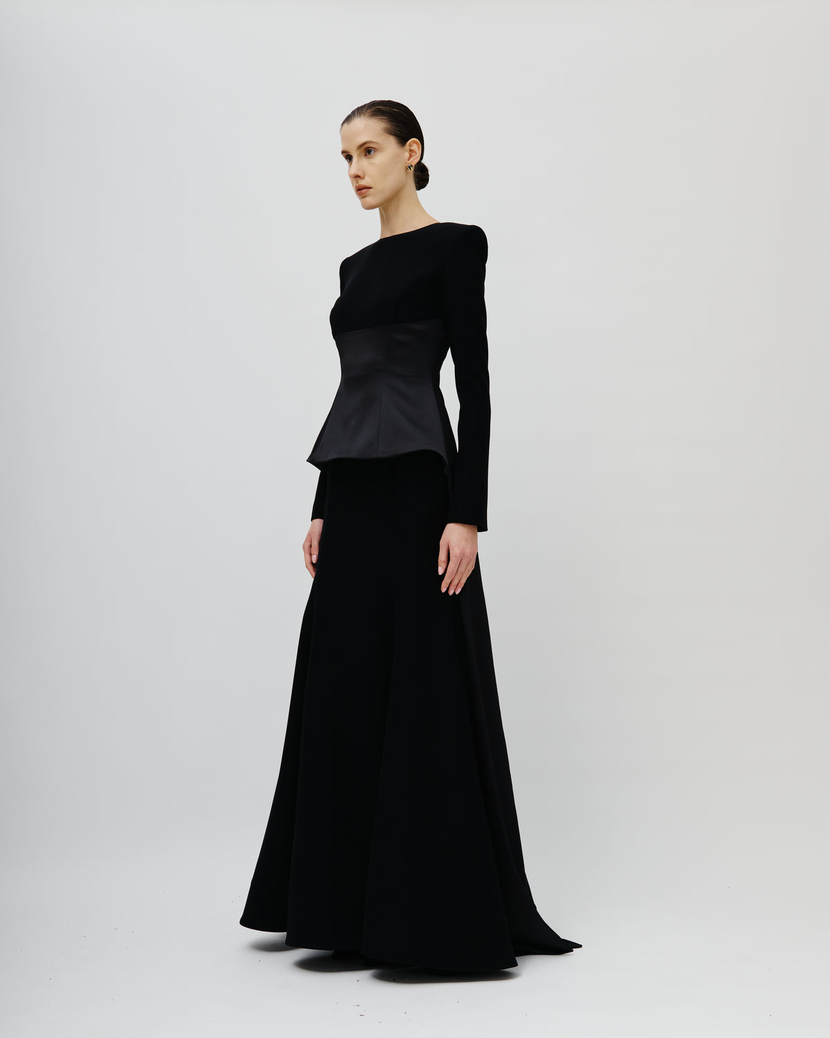 A model wearing a long black evening Avaro Figlio gown with structured shoulders, a peplum waist, and a flowing skirt, photographed against a seamless light gray studio background.